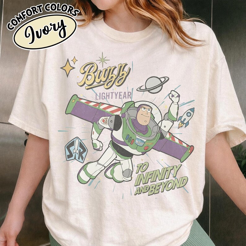 Może przedstawiać: Kremowa koszulka z grafiką Buzz'a Lightyeara w pozie lotu. Projekt zawiera tekst "Buzz Lightyear" i "To Infinity and Beyond" z rakietą i ilustracją planety. Koszulka jest z Comfort Colors.