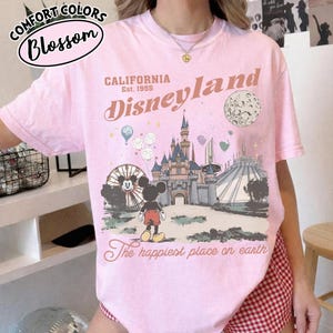 May include: Pink t-shirt with a Disneyland graphic. The design includes the Disneyland logo, a castle, Mickey Mouse, a Ferris wheel, and the text "The happiest place on earth." The shirt also has the text "Comfort Colors Blossom" at the top.