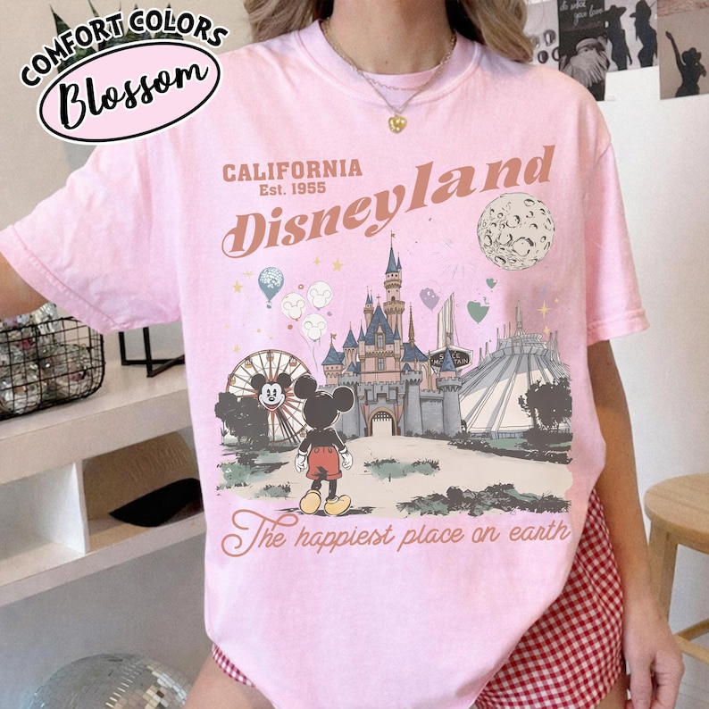 May include: Pink t-shirt with a Disneyland graphic. The design includes the Disneyland logo, a castle, Mickey Mouse, a Ferris wheel, and the text "The happiest place on earth." The shirt also has the text "Comfort Colors Blossom" at the top.