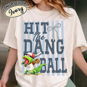 Puede incluir: Camiseta color marfil con el texto azul "HIT the DANG BALL". Un lagarto de dibujos animados con uniforme de béisbol sostiene un bate. La camiseta también presenta un lazo y rayas azules verticales.