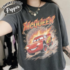 May include: Dark gray t-shirt featuring a graphic of the character Lightning McQueen from the movie Cars. The design includes the name "McQueen" in orange and red flames, and the number "95" on the car. The shirt has the text "Comfort Colors Pepper" in a white oval.
