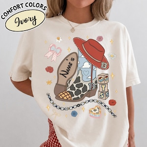 Puede incluir: Una camiseta color marfil con un gr&aacute;fico de una bota vaquera con estampado de vaca, un sombrero rojo y bloques de letras. El dise&ntilde;o incluye el texto "NAME'S", "13" y "I KNEW IT I KNEW YOU" en un cord&oacute;n de cuentas, rodeado de flores y estrellas.
