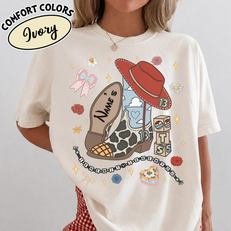 Puede incluir: Una camiseta color marfil con un gr&aacute;fico de una bota vaquera con estampado de vaca, un sombrero rojo y bloques de letras. El dise&ntilde;o incluye el texto "NAME'S", "13" y "I KNEW IT I KNEW YOU" en un cord&oacute;n de cuentas, rodeado de flores y estrellas.