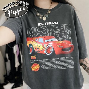 May include: Dark gray t-shirt featuring a graphic of the character Lightning McQueen from the movie Cars. The design includes the text "EL RAYO MCQUEEN" and "95". The shirt also has the "Rust-eze" logo.