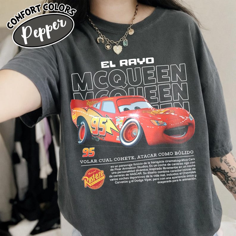 May include: Dark gray t-shirt featuring a graphic of the character Lightning McQueen from the movie Cars. The design includes the text "EL RAYO MCQUEEN" and "95". The shirt also has the "Rust-eze" logo.