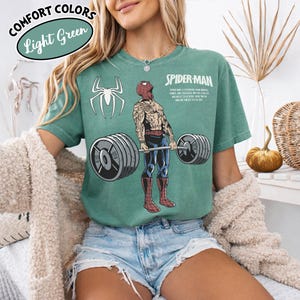 May include: Light green t-shirt with a Spider-Man graphic lifting weights. The shirt features the text "COMFORT COLORS" and "Light Green" in a circular design. A white spider logo is on the left side of the shirt.