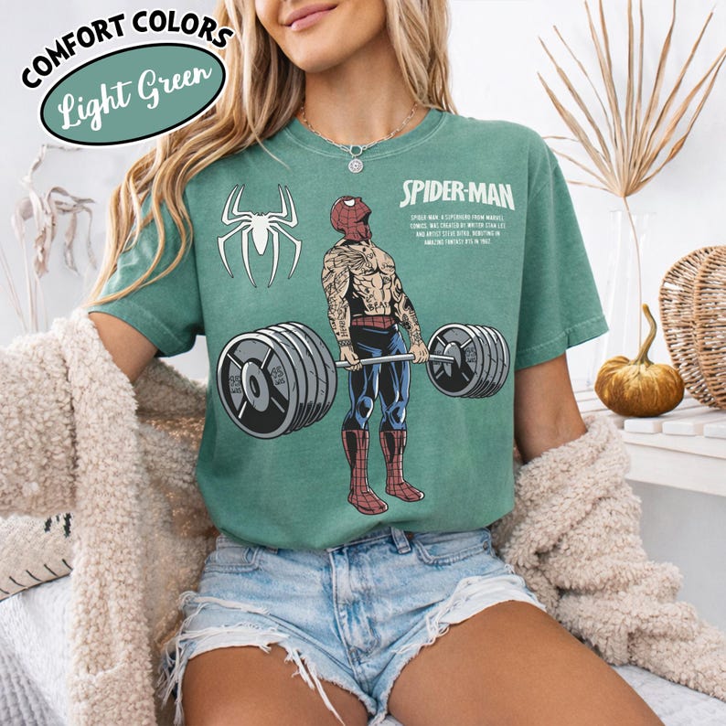 May include: Light green t-shirt with a Spider-Man graphic lifting weights. The shirt features the text "COMFORT COLORS" and "Light Green" in a circular design. A white spider logo is on the left side of the shirt.