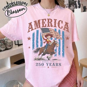 Puede incluir: Una camiseta de manga corta de color rosa claro con un gr&aacute;fico de una vaquera montando un caballo mientras sostiene una bandera estadounidense. El texto dice AMERICA, SINCE 1776 y 250 YEARS. Incluye rayas verticales azules y estrellas rojas.