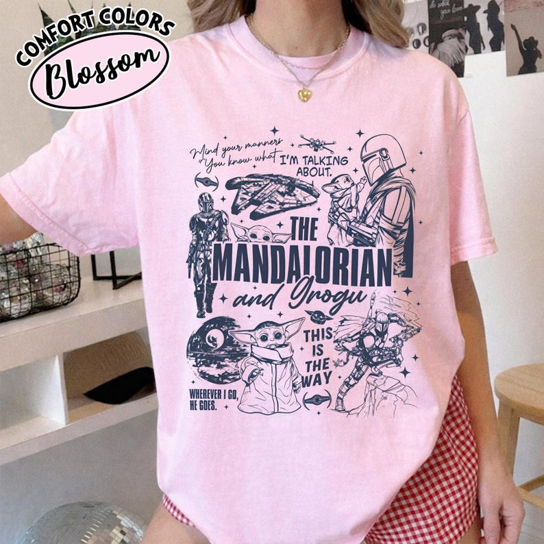 Op de afbeelding: Lichtroze t-shirt met een grafisch ontwerp van personages en elementen uit de serie "The Mandalorian". Het ontwerp bevat de tekst "The Mandalorian and Grogu" en andere zinnen. Het t-shirt heeft korte mouwen.