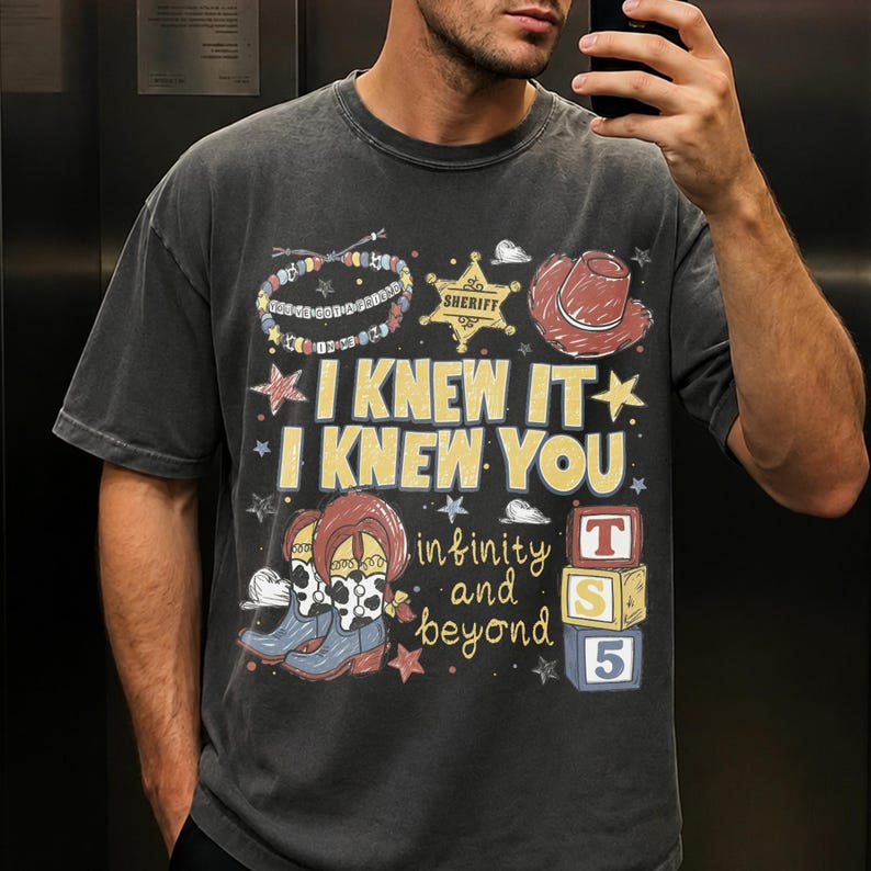May include: A charcoal grey short-sleeve t-shirt featuring a graphic print with the text I KNEW IT I KNEW YOU, infinity and beyond, and TS 5. The design includes a beaded bracelet, a sheriff star badge, a red cowboy hat, and cow-print boots.