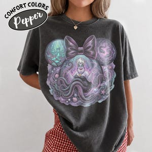 May include: Dark gray t-shirt featuring a graphic of Ursula from The Little Mermaid, with a large bow and Mickey Mouse ears. The design includes the text "COMFORT COLORS Pepper". The shirt is a comfortable fit.
