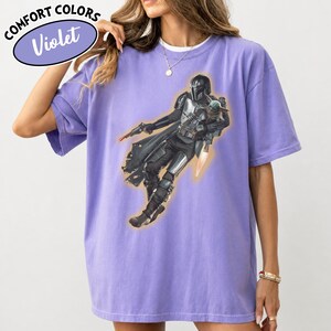 Puede incluir: Una camiseta violeta de manga corta con un estampado gr&aacute;fico de un personaje con armadura y casco que sostiene a una peque&ntilde;a criatura verde. El personaje lleva una armadura met&aacute;lica oscura, una capa, un bl&aacute;ster y un jetpack con llamas.