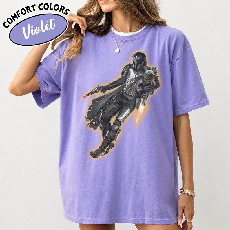 Puede incluir: Una camiseta violeta de manga corta con un estampado gr&aacute;fico de un personaje con armadura y casco que sostiene a una peque&ntilde;a criatura verde. El personaje lleva una armadura met&aacute;lica oscura, una capa, un bl&aacute;ster y un jetpack con llamas.