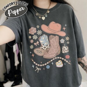 May include: A charcoal gray t-shirt featuring a graphic print of a peach cowboy hat, two cowboy boots, flowers, stars, a heart-shaped cake, and a beaded string with the text "I KNEW IT I KNEW YOU". The shirt has a crew neck and short sleeves.