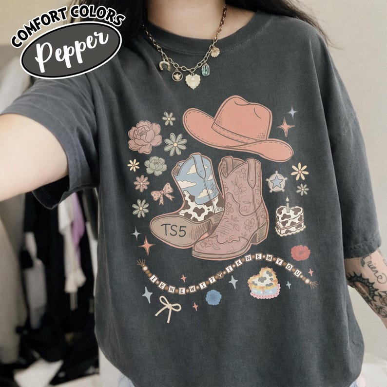May include: A charcoal gray t-shirt featuring a graphic print of a peach cowboy hat, two cowboy boots, flowers, stars, a heart-shaped cake, and a beaded string with the text "I KNEW IT I KNEW YOU". The shirt has a crew neck and short sleeves.