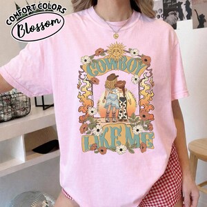 May include: A light pink t-shirt featuring a graphic print of two figures in cowboy hats and boots standing under a sun and floral arch. The text COWBOY LIKE ME is printed in large blue letters. The design includes orange flames and white flowers.