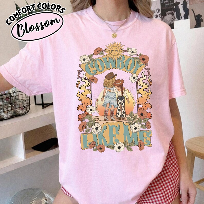 May include: A light pink t-shirt featuring a graphic print of two figures in cowboy hats and boots standing under a sun and floral arch. The text COWBOY LIKE ME is printed in large blue letters. The design includes orange flames and white flowers.