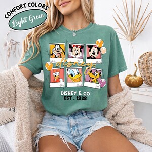 Op de afbeelding: Lichtgroen T-shirt met een Disney-ontwerp. Het shirt toont zes karakterportretten, waaronder Mickey Mouse, Minnie Mouse, Donald Duck en Goofy. De tekst "Disney & Co Est. 1928" staat eronder gedrukt. Het shirt heeft ook de tekst "Comfort Colors Light Green".
