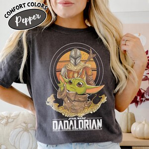 Może przedstawiać: Ciemnoszara koszulka z grafiką The Mandalorian i Grogu z Gwiezdnych Wojen. Projekt zawiera okrągłą ramkę z tłem zachodu słońca. Tekst "STAR WARS DADALORIAN" jest wydrukowany na dole. Koszulka ma u g&oacute;ry napis "COMFORT COLORS Pepper".