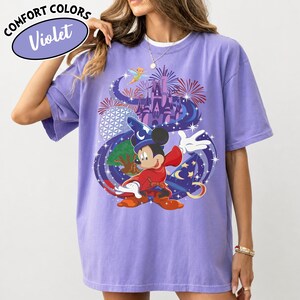 Może przedstawiać: Fioletowa koszulka Comfort Colors z grafiką Myszki Miki w stroju czarodzieja, zamkiem, fajerwerkami i Dzwoneczkiem. Koszulka ma napis "Comfort Colors" i "Violet" w okrągłym wzorze.
