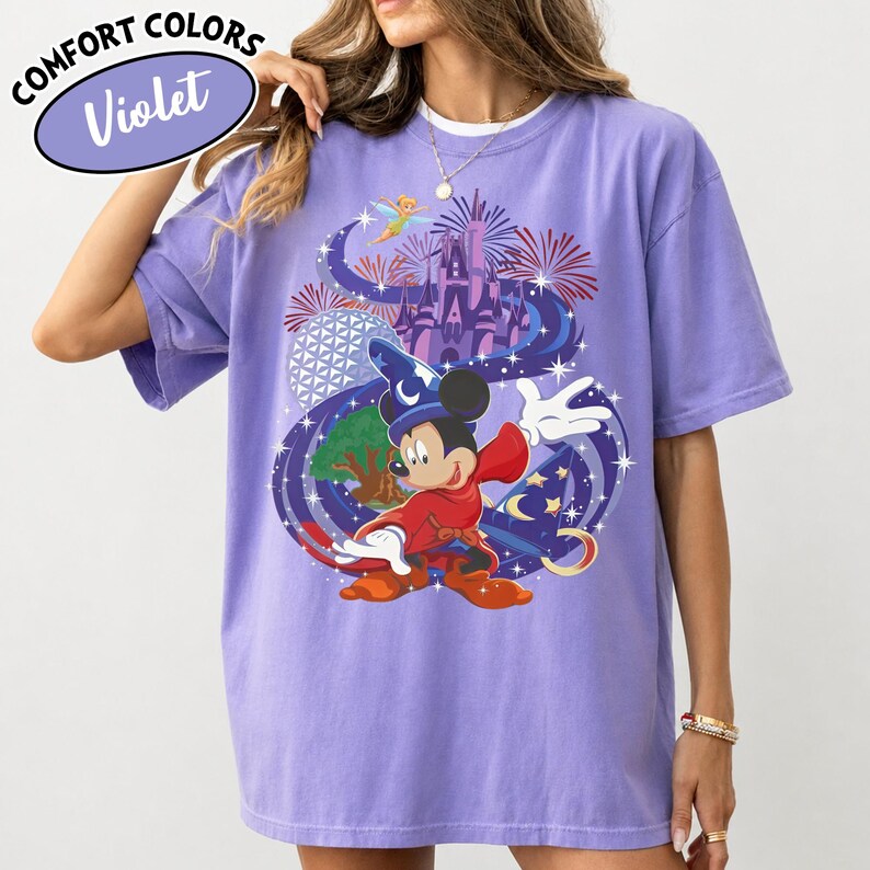Może przedstawiać: Fioletowa koszulka Comfort Colors z grafiką Myszki Miki w stroju czarodzieja, zamkiem, fajerwerkami i Dzwoneczkiem. Koszulka ma napis "Comfort Colors" i "Violet" w okrągłym wzorze.