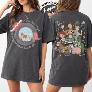 May include: A charcoal gray short-sleeve t-shirt featuring a front graphic of a circular frame with a cowboy hat and boot, text reading Love has ways of bringing things back to life, and a back graphic of various animated characters with names.