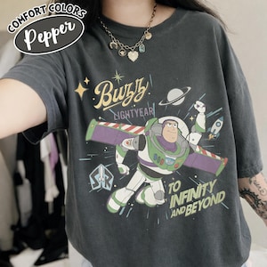 Może przedstawiać: Ciemnoszara koszulka z Buzzem Astralem z Toy Story. Grafika zawiera tekst "Buzz Lightyear" i "To Infinity and Beyond" z motywem kosmicznym. Koszulka ma etykietę Comfort Colors Pepper.