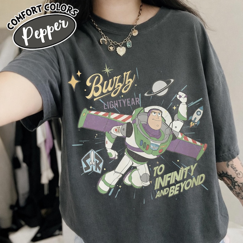Może przedstawiać: Ciemnoszara koszulka z Buzzem Astralem z Toy Story. Grafika zawiera tekst "Buzz Lightyear" i "To Infinity and Beyond" z motywem kosmicznym. Koszulka ma etykietę Comfort Colors Pepper.