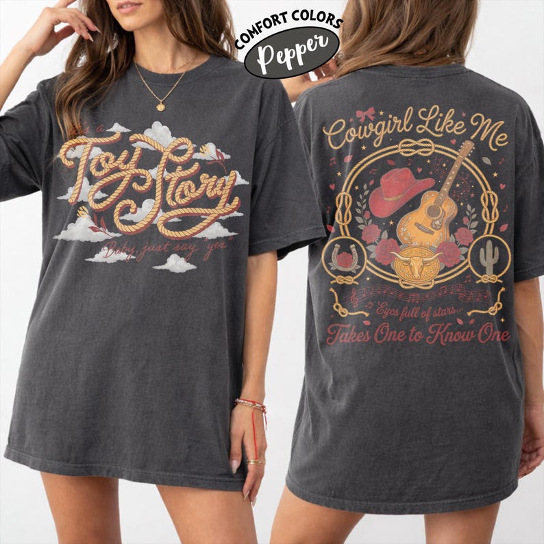 May include: A charcoal gray short-sleeve t-shirt featuring a front graphic with the text Toy Story in a rope-style font over clouds. The back graphic displays the text Cowgirl Like Me and Takes One to Know One with a guitar, hat, and bull skull.