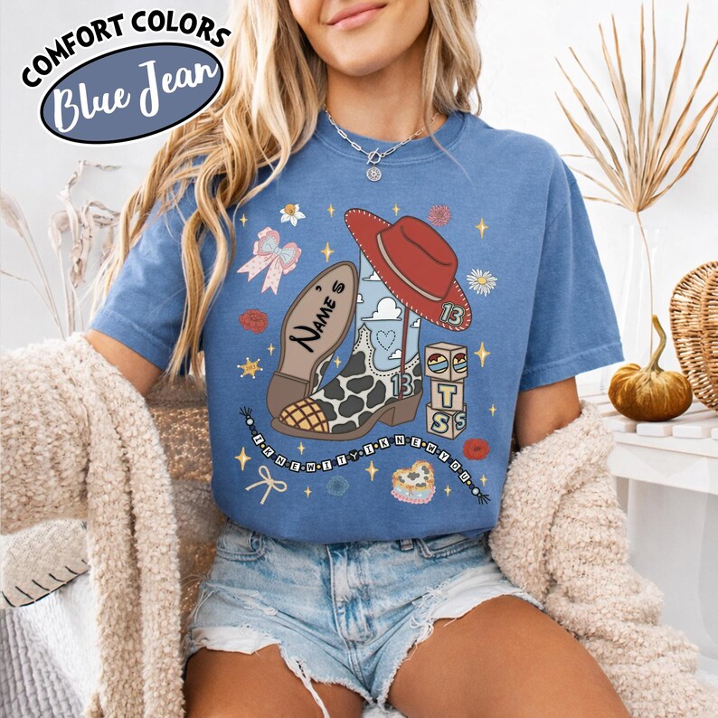 Puede incluir: Una camiseta azul con un estampado gr&aacute;fico de una bota de vaquero con patr&oacute;n de vaca, un sombrero de vaquero rojo, un lazo rosa y bloques apilados con las letras T, S y S. El texto "Name's" est&aacute; en la bota y "13" en el sombrero.