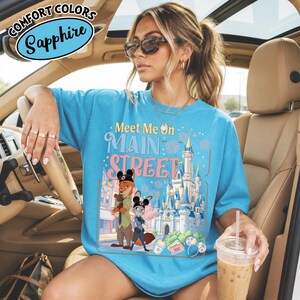 May include: Sapphire blue t-shirt with the text "Meet Me On Main Street" and a graphic of cartoon characters and a castle. The shirt is a Comfort Colors brand.