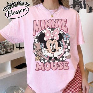 May include: Pink t-shirt featuring a Minnie Mouse graphic. The design includes Minnie's face, a polka dot bow, and the words "MINNIE MOUSE". The shirt also has the text "COMFORT COLORS Blossom" in a circular design.