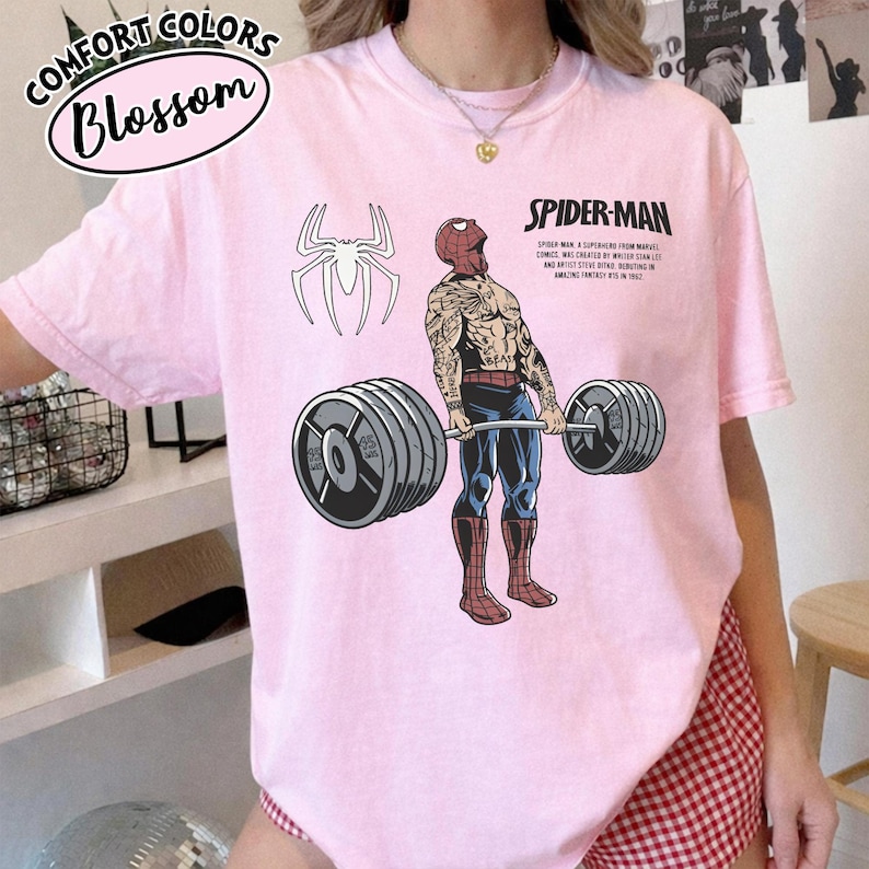 May include: A light pink t-shirt featuring a Spider-Man graphic lifting weights. The design includes a white spider symbol, the words "Spider-Man", and text describing the character's origin. The shirt also has the words "Comfort Colors Blossom" in a circular design.