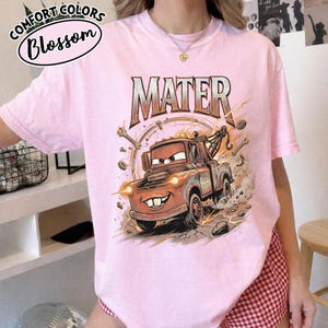 Puede incluir: Camiseta rosa claro con un dise&ntilde;o de Mate de la pel&iacute;cula Cars. El dise&ntilde;o incluye la palabra "MATER" en negrita sobre el personaje. La camiseta tambi&eacute;n tiene las palabras "Comfort Colors" y "Blossom" impresas en la parte superior izquierda.