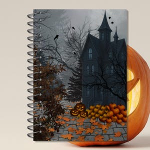 Puede incluir: Un cuaderno con espiral con una portada de Halloween. La portada muestra una casa embrujada, calabazas y hojas de otoño. Una calabaza tallada con una cara brillante está en el lado derecho.