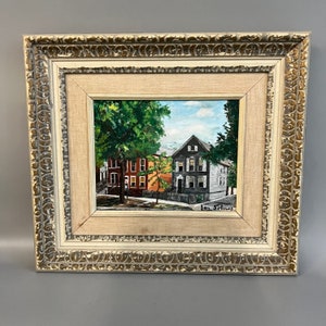 May include: Framed painting of a street scene with three houses. The houses are painted in shades of gray, red, and orange. The painting is signed "Jon Simes" in the bottom right corner.