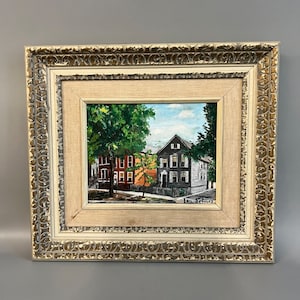 May include: A framed oil painting depicting a street scene with two houses. The house on the left is red brick with white trim, and the house on the right is gray with white trim. There is a large green tree in the foreground. The painting is signed "Ann St. James".