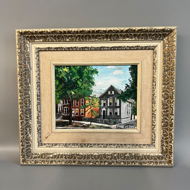 May include: A framed oil painting depicting a street scene with two houses. The house on the left is red brick with white trim, and the house on the right is gray with white trim. There is a large green tree in the foreground. The painting is signed "Ann St. James".