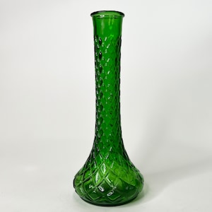 May include: A green glass vase with a diamond pattern. The vase has a flared base and a slender, cylindrical neck. The green colour is vibrant, and the glass appears textured. The vase is likely used for displaying flowers. The height is approximately 25 cm.