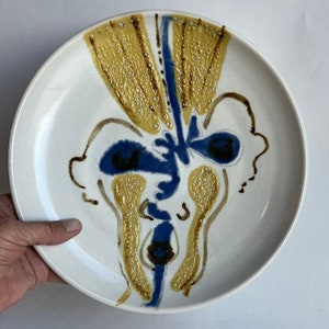 May include: A white ceramic plate with a blue and yellow abstract design. The design features a face with two eyes, a nose, and a mouth.