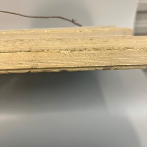 May include: Close-up of the bottom edge of a light beige wooden frame. The frame has a slightly distressed look with visible wood grain.
