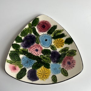 May include: A white ceramic triangular plate with a black rim and a floral design. The design features various flowers in shades of pink, blue, yellow, and purple, with green leaves.