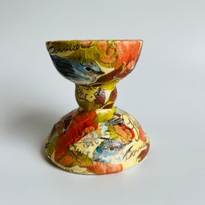May include: A decorative ceramic egg cup with a two-tiered design. The cup is decorated with a collage of vintage paper featuring birds, flowers, and text. The colors are predominantly green, yellow, orange, and blue.