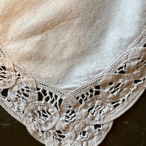 May include: A close-up of a white linen tablecloth with a delicate lace trim. The lace is made of small, intricate loops and knots, creating a delicate and elegant design.