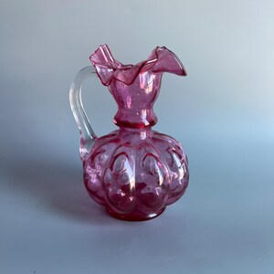 May include: A pink glass pitcher with a ruffled top and a clear glass handle. The pitcher has a textured, embossed design.