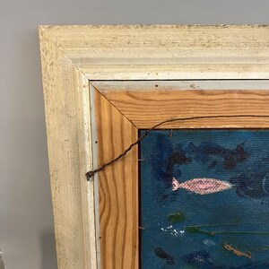 May include: A close-up of the top corner of a framed painting. The frame is made of wood and painted white. The painting is on a blue canvas and features a pink fish.