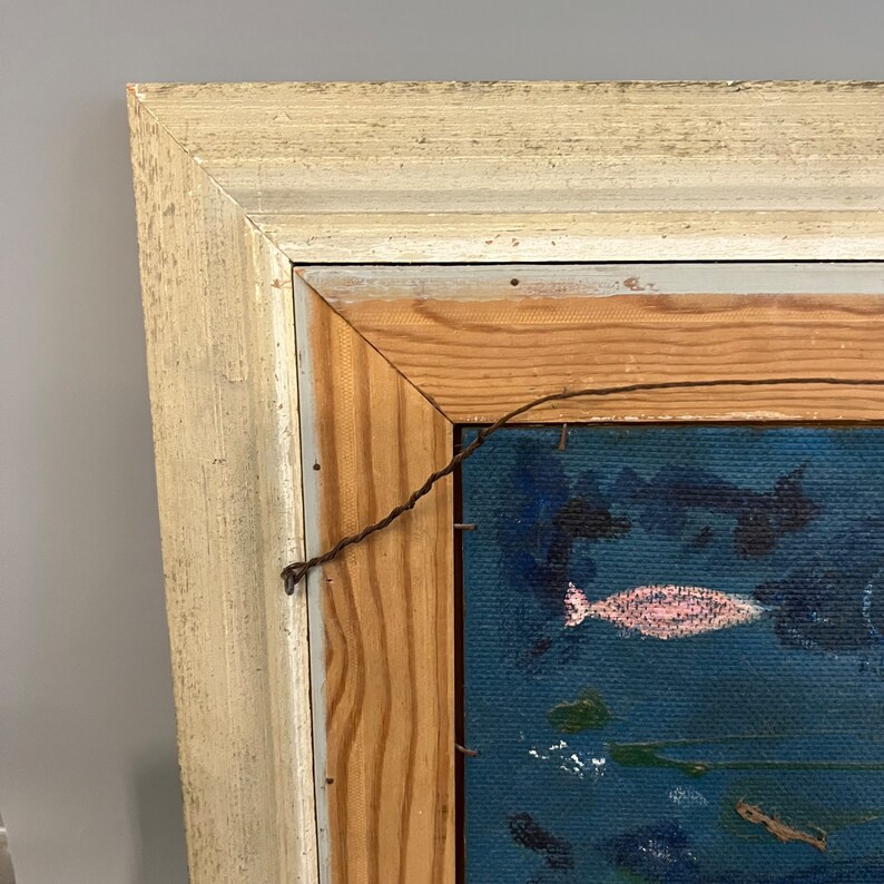 May include: A close-up of the top corner of a framed painting. The frame is made of wood and painted white. The painting is on a blue canvas and features a pink fish.