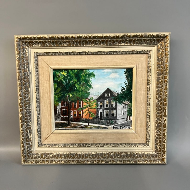 May include: A framed oil painting depicting a street scene with two houses. The house on the left is orange and the house on the right is gray. There are trees in the foreground and a blue sky in the background. The painting is signed "Jan Selmes".