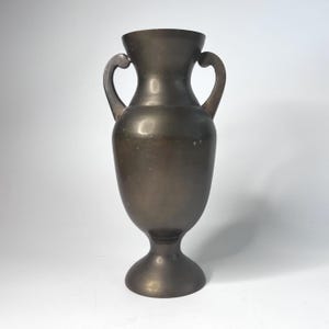 May include: Bronze-colored vase with two curved handles. The vase has a wide base and a flared top. The overall shape is symmetrical and elegant, suitable for floral arrangements or as a decorative piece. The vase is made of metal.