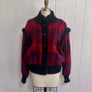 May include: A vintage red and purple plaid cardigan with black accents. The button-up sweater features a high collar, black buttons, and black trim on the sleeves and hem. The sweater is displayed on a mannequin.